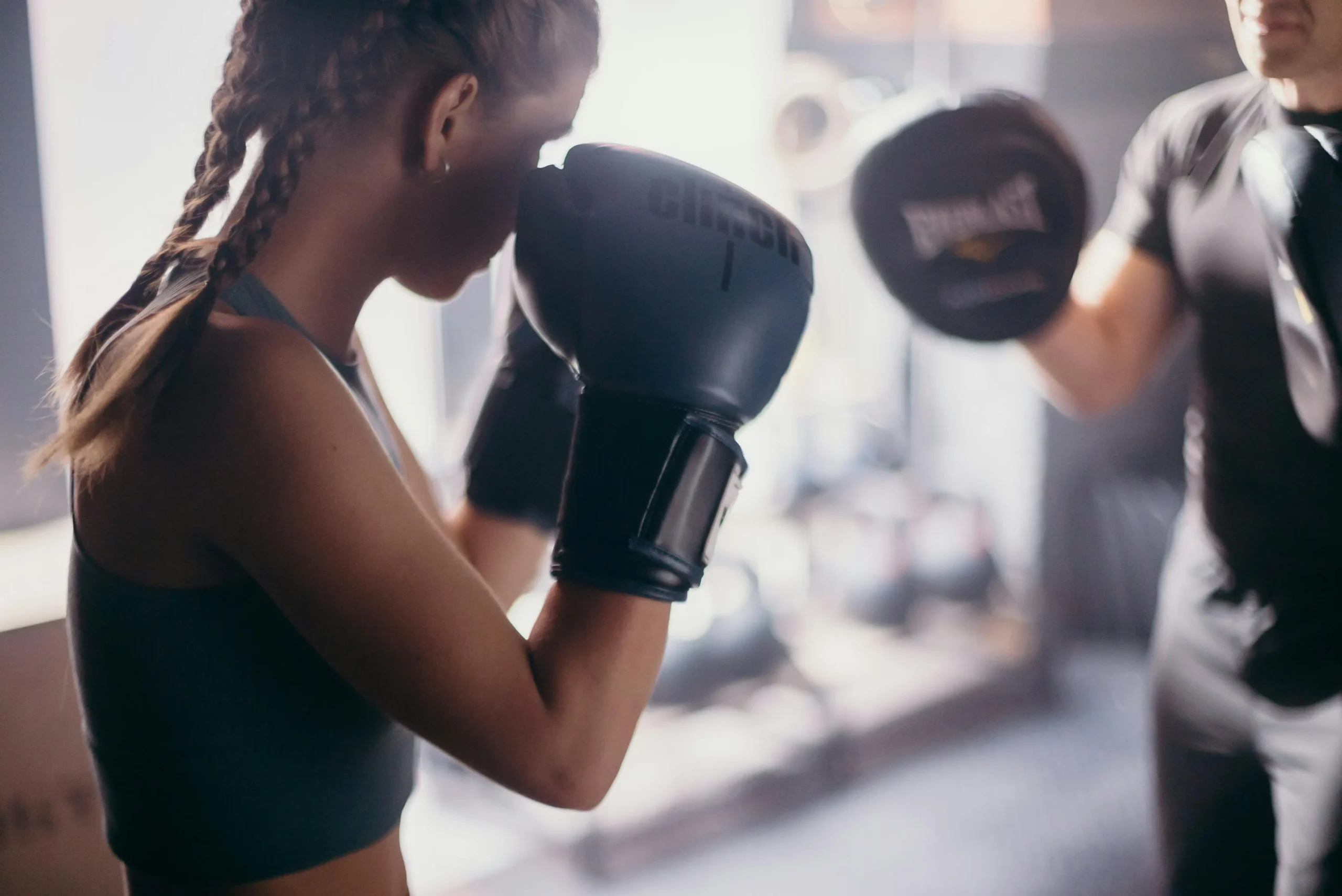 A woman in boxing gloves stands confidently, with a man training in the background.<br />

